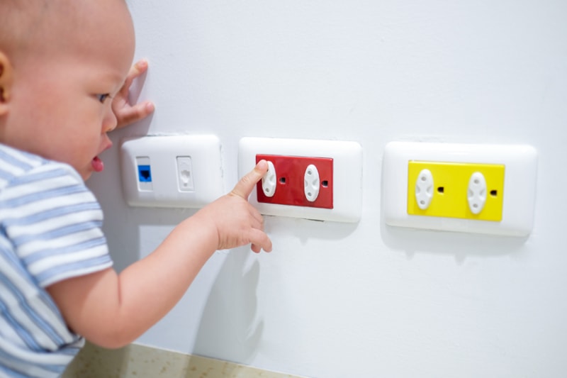 Curious toddler baby boy child play with electric plug. Try to insert it into socket, Baby hand reach for electrical outlet covered with red safety plugs (child safety concept) - Selective focus.